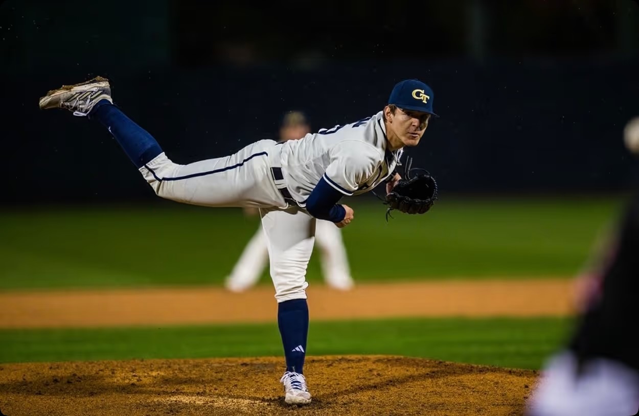 A college pitcher at full extension on the mound during a night game.